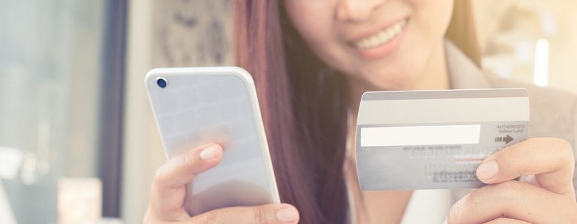 Une femme souriante tenant sa carte bancaire et son smartphone pour faire un achat en ligne