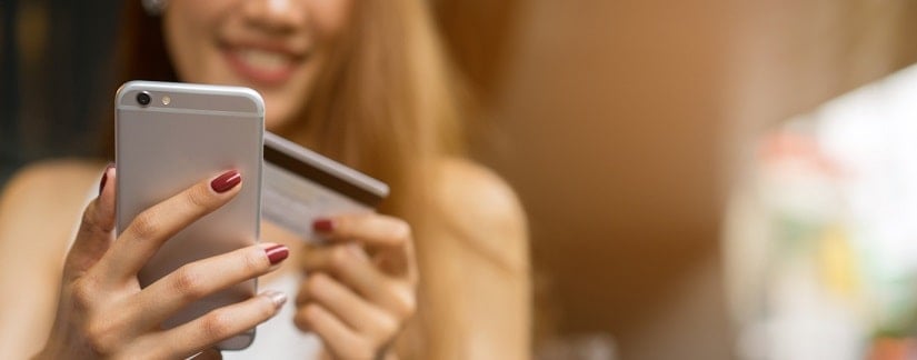 Une souriante jeune femme utilise son téléphone en tenant sa carte bancaire pour faire un achat de produits en ligne.