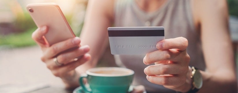 Main d’une femme au café en tenant sa carte bancaire et son téléphone pour faire des achats en ligne.