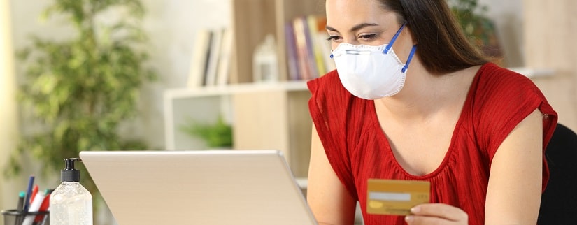 Femme payant en ligne avec sa carte sur l’ordinateur portable portant un masque en confinement Covid-19 assis sur un bureau à la maison.