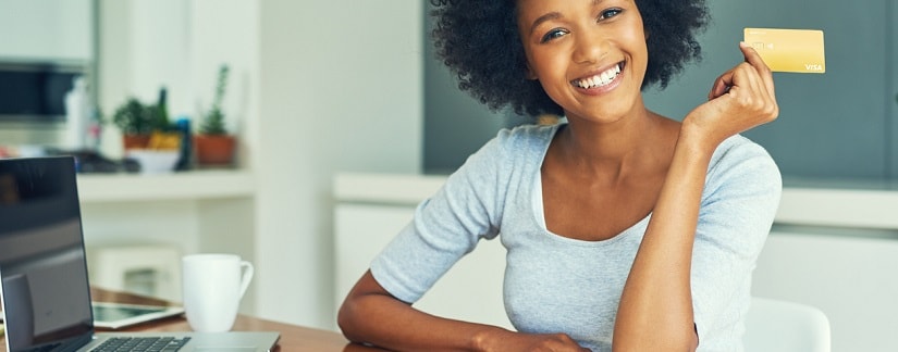 Une jeune Africaine souriante au bureau en tenant sa carte de crédit sur l’ordinateur portable faisant ses opérations bancaires en ligne.