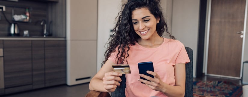 Une jeune femme se détendre à la maison en tenant sa carte bancaire et son smartphone pour faire des achats en ligne.
