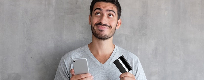 Jeune homme beau penser à des achats en ligne via Internet, portant t-shirt gris, debout contre le mur texturé, tenant la carte de crédit
