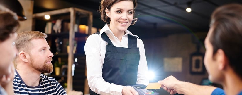 Heureuse jeune serveuse positive avec les cheveux courts debout à table et tenant lecteur de carte