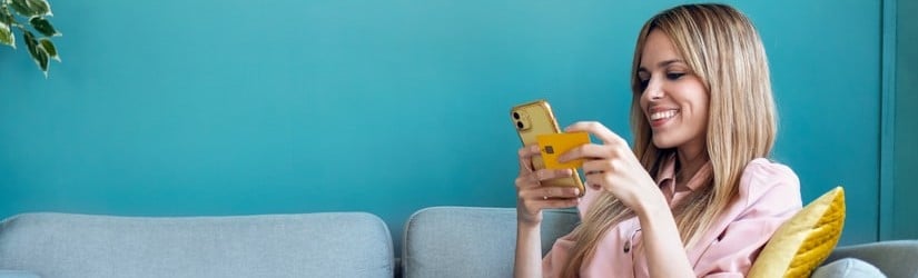 Une femme assise sur un canapé tenant un smartphone et un crédit carte pour faire des services en ligne.