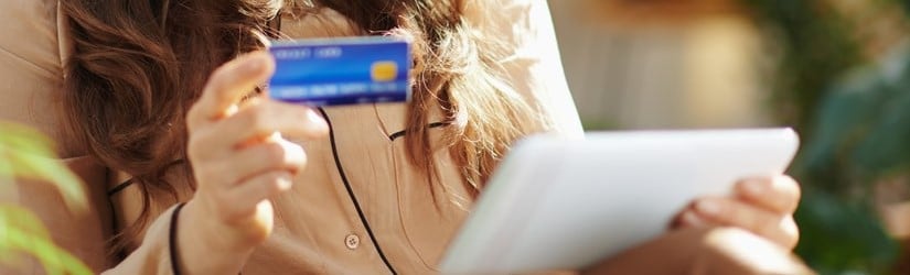 Femme moderne heureuse aux longs cheveux ondulés avec une carte de crédit utilisant une tablette PC dans une maison moderne en plein soleil.