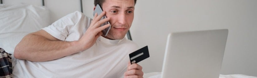 Jeune homme en chemise blanche et pyjama assis sur un lit regardant l'écran tenant une carte bancaire.