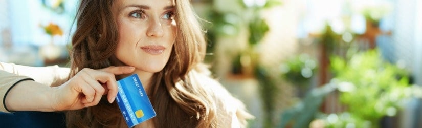 Femme au foyer pensive et élégante aux longs cheveux ondulés avec une carte de crédit faisant des achats en ligne sur un site de commerce électronique dans une maison moderne en plein soleil.
