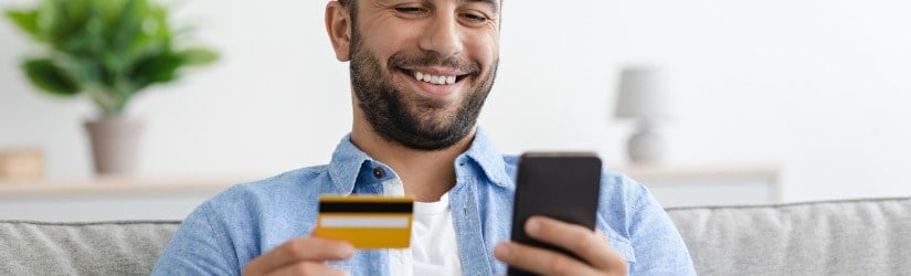 Un homme adulte caucasien souriant et satisfait avec une barbe tient une carte de crédit et paie de l'argent en ligne sur un smartphone dans un intérieur de maison, espace libre. 