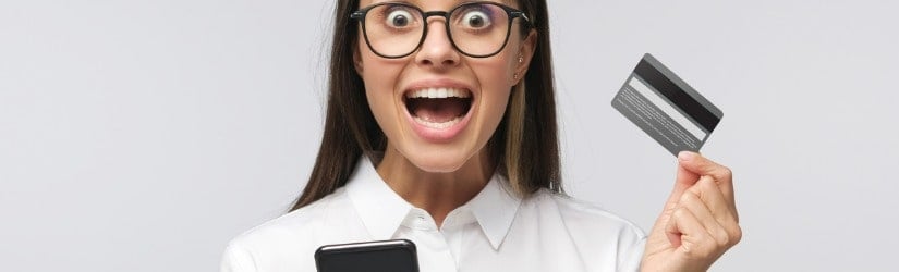 Portrait d'une jeune femme excitée, bouche ouverte, tenant une carte de crédit et utilisant un téléphone portable.