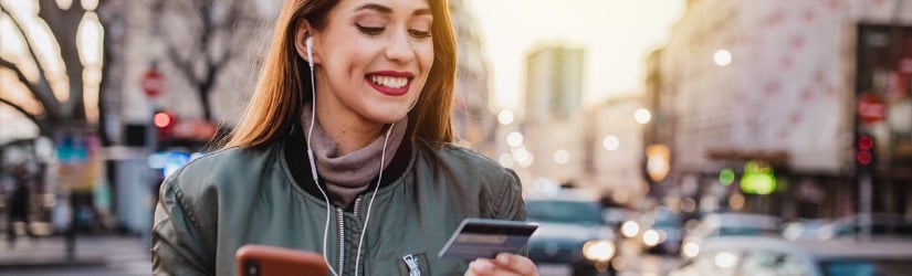 Une jeune femme heureuse avec un téléphone portable fait des achats en ligne avec sa carte de crédit et son smartphone à l'extérieur.