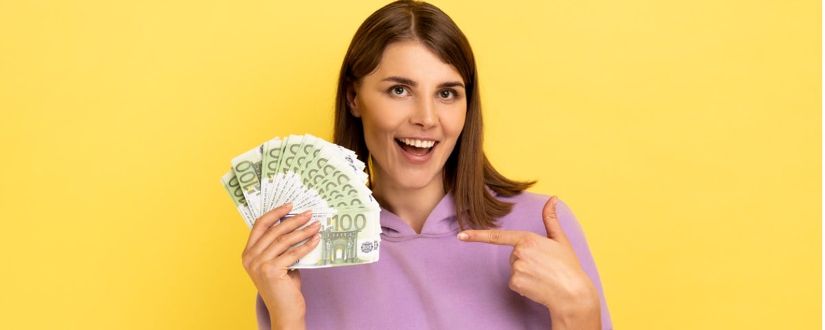 Femme souriante et stupéfaite montrant des billets de banque en euros dans sa main, ayant une expression positive optimiste, un gros bénéfice, portant un sweat à capuche violet. 