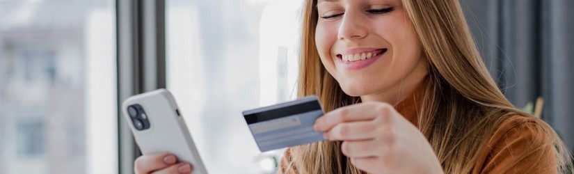Jeune femme assise à une table et faisant des achats en ligne elle tient un téléphone portable et une carte de crédit. 