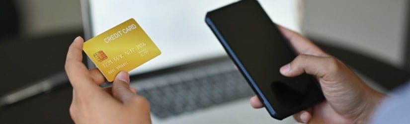 Image recadrée d'un jeune homme d'affaires tenant une carte de crédit et un smartphone pour effectuer un paiement en ligne devant un ordinateur portable au bureau.