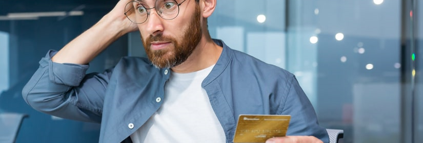 Jeune homme choqué, assis dans un bureau, carte de crédit en main, ordinateur portable en main. 